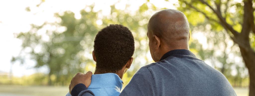 A man comforting his son with a hand on his shoulder outside in a park
