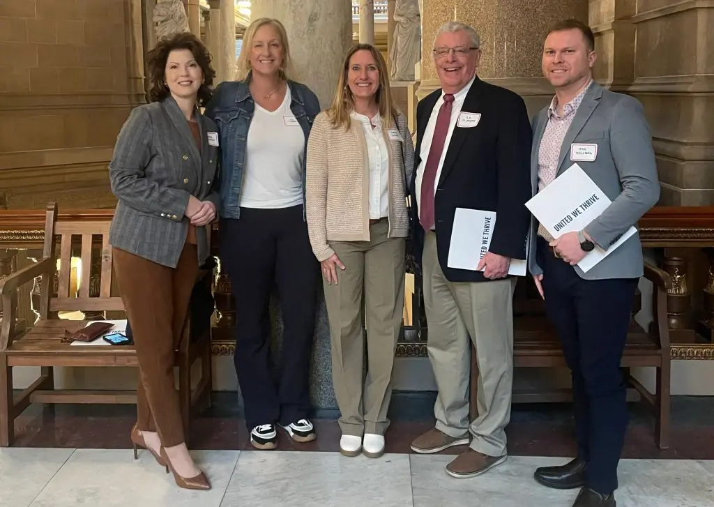 ive adults standing together inside a government building, some holding folders labeled “United We Thrive.”