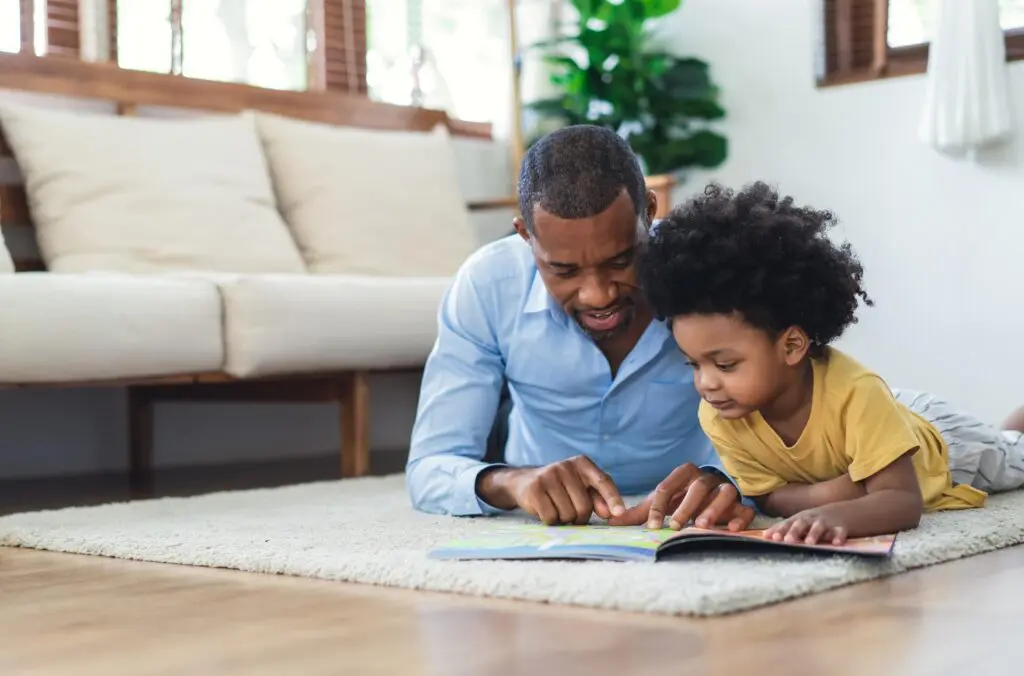 Happy African American father and son are reading a book and smiling while lying on floor spending time together at home.