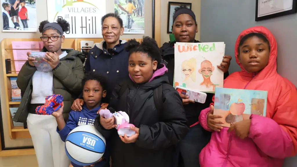 A woman stands with five children indoors as several of the children hold toys, a basketball and colorful artwork.