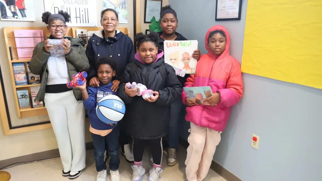 Group of children and an adult standing indoors by a wall display, holding toys, a basketball, and a drawing labeled “Wicked.”