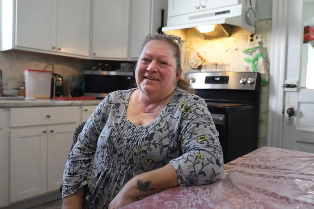 A woman in a floral blouse sits at a kitchen table, smiling. The kitchen has white cabinets, a stove, and a microwave in the background.