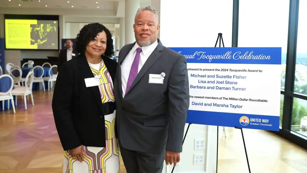 Two people standing in front of a sign at the Annual Tocqueville Celebration, which recognizes significant contributors to the United Way in Greater Cincinnati. The sign lists the 2021 Tocqueville Award recipients and the newest members of the Million Dollar Roundtable.