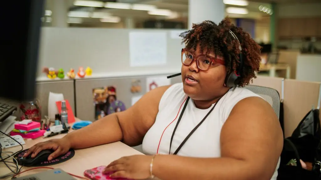 Person wearing a headset working at a desk in an office cubicle, with a computer, soda can, papers, and personal decorations on the cubicle walls.