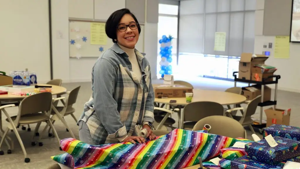 Person smiling while wrapping gifts in colorful paper at a table in a room with chairs, boxes, and decorations.