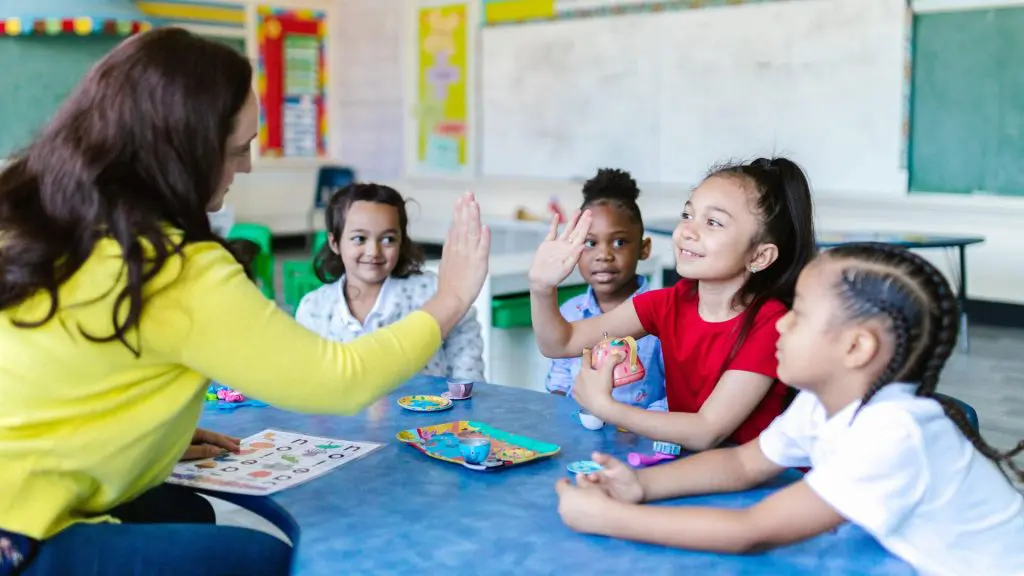 Teacher in a yellow sweater giving high-fives to two children at a blue table in a classroom, with colorful toys and playdough on the table and educational posters in the background.