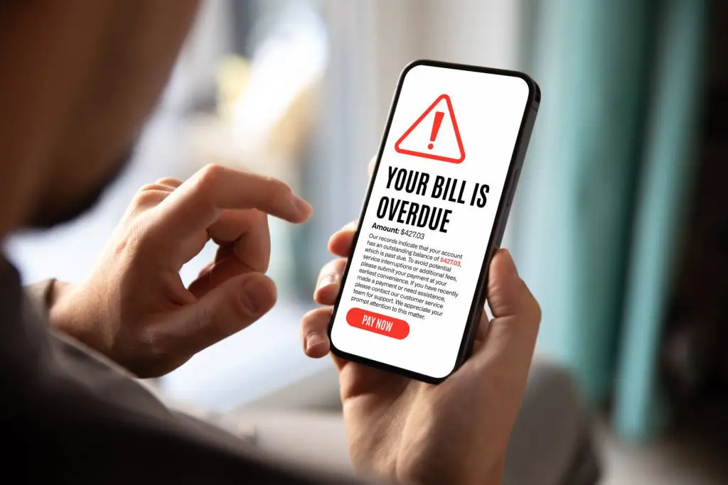 A person holding a smartphone displaying an overdue bill notification for $427.03, with a prompt to pay now to avoid service interruption.