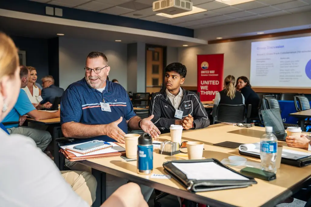 Group of people seated around a table during a meeting, with notebooks, coffee cups, and folders visible. A presentation slide in the background displays 'United Way' and 'UNITED WE THRIVE.'