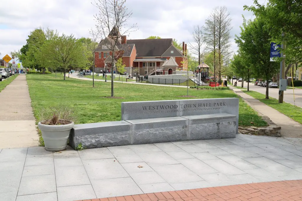 Stone bench engraved with 'WESTWOOD TOWN HALL PARK' in a grassy park area, with a red brick building and trees in the background under a partly cloudy sky.