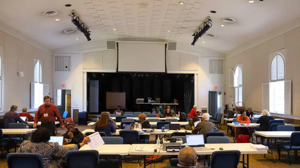 People sit at tables with laptops and paperwork inside a community center during a free tax preparation event. A stage with a large projection screen is visible at the front of the room as volunteers assist attendees throughout the space.