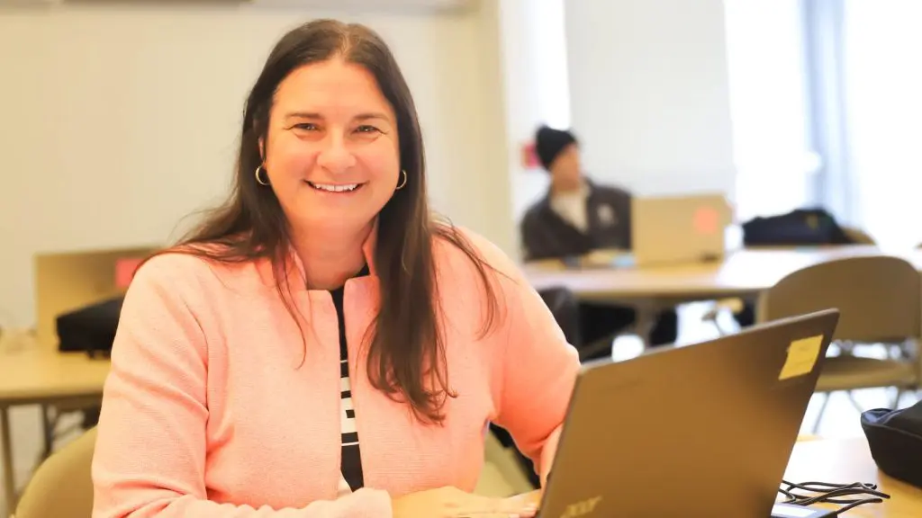 Smiling person in a pink jacket sitting at a table with a laptop in a classroom or meeting room.