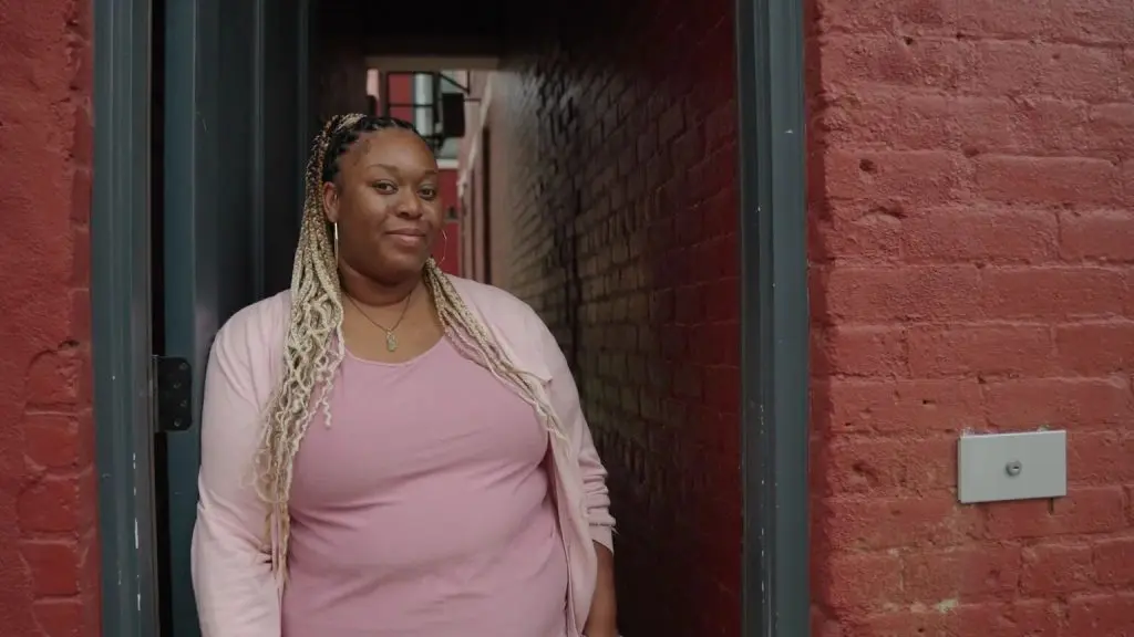 Person with long braided hair wearing a pink top and cardigan standing in a doorway between red brick walls.