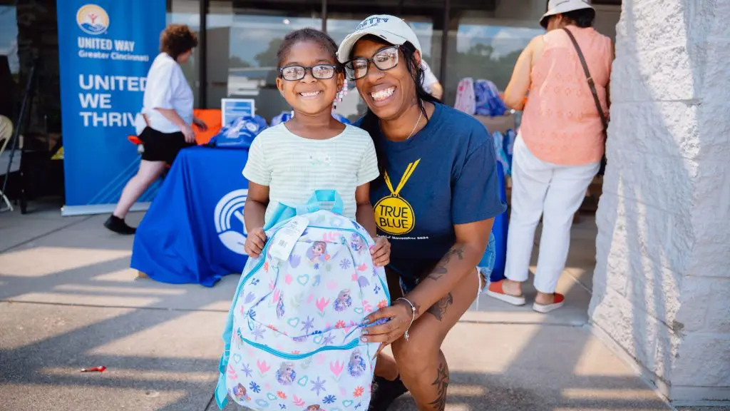 A young girl in glasses smiles holding a Frozen-themed backpack with a woman kneeling beside her at a United Way of Greater Cincinnati backpack giveaway event