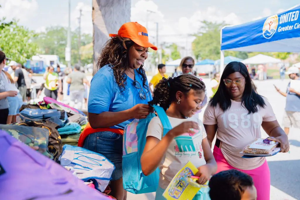 Volunteers and families at an outdoor event with a United Way of Greater Cincinnati tent, distributing backpacks and school supplies to children.