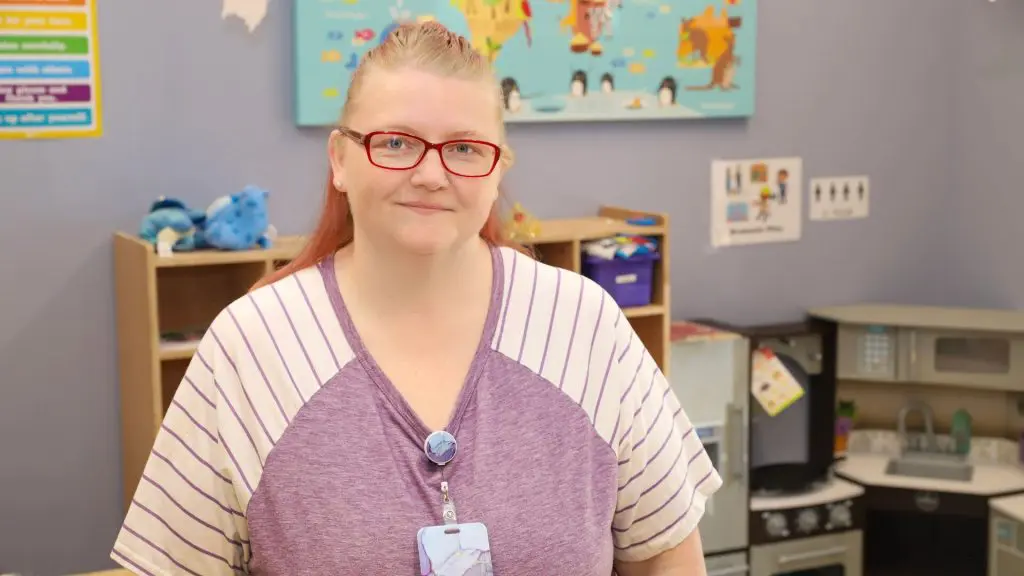 A person wearing red glasses and a purple shirt stands in a childcare classroom, smiling at the camera. Shelves with toys and educational materials, a play kitchen, and a colorful world map are visible in the softly blurred background.