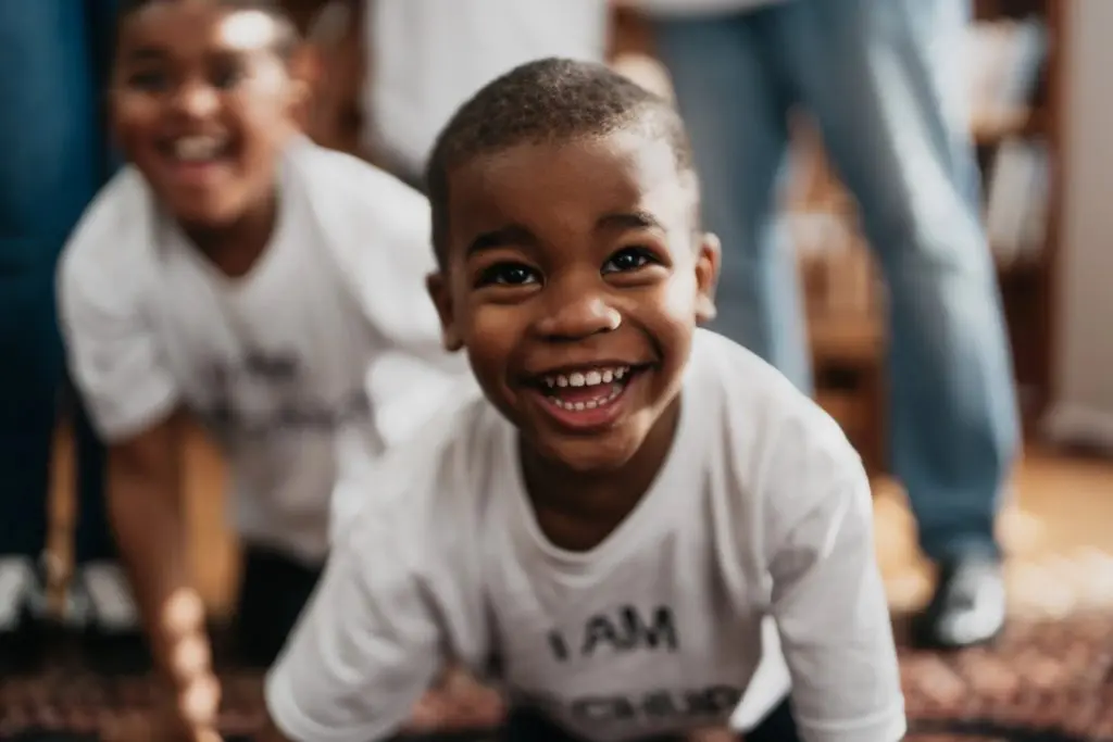 Child smiling and leaning forward toward the camera, with another child visible in the background.