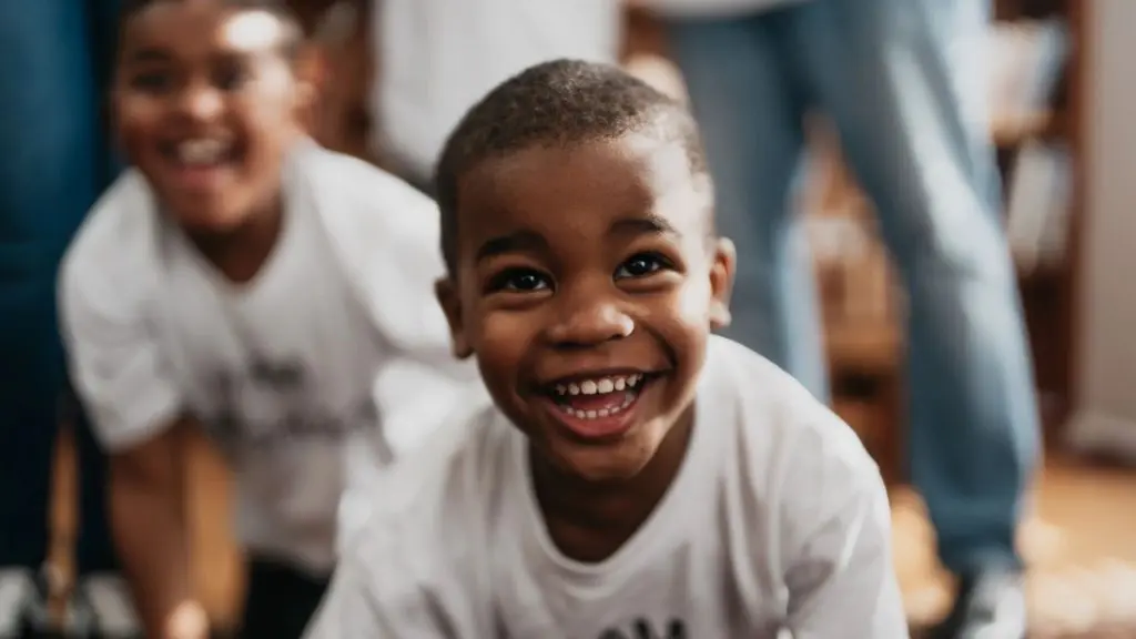 Child smiling brightly while leaning toward the camera, with another child blurred in the background.