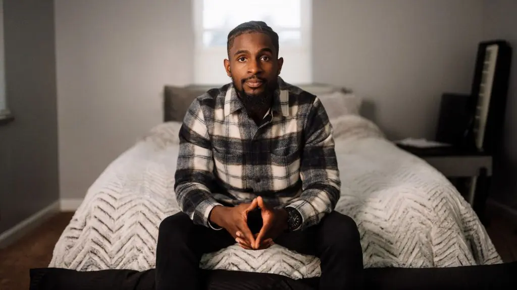 Person sitting on the edge of a neatly made bed, looking at the camera in a softly lit bedroom.