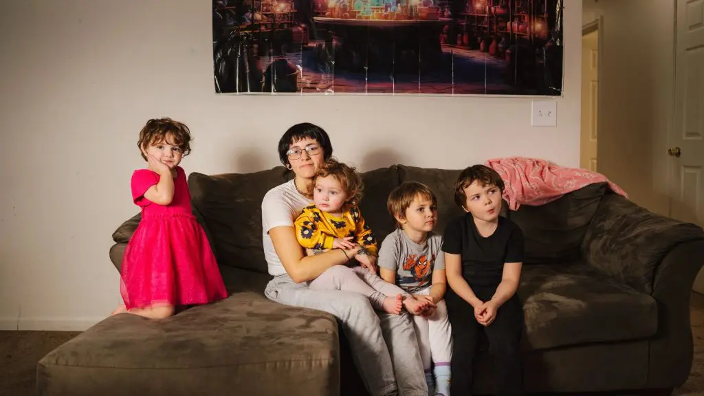 Adult sitting on a couch holding a young child, with three other children nearby, all in a living room with a large poster on the wall.