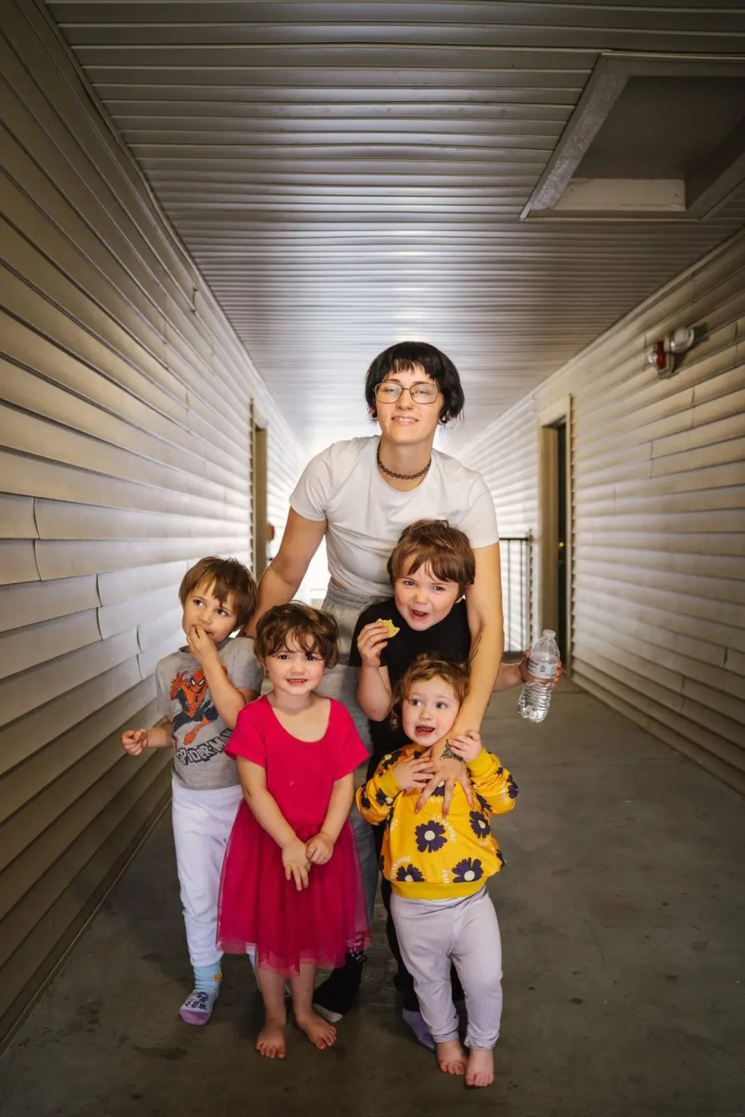Adult standing in an outdoor hallway with four young children gathered close and smiling toward the camera.