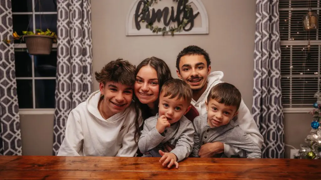 Family of five smiling closely together at a wooden table in a home, with patterned curtains and a “family” sign on the wall behind them.