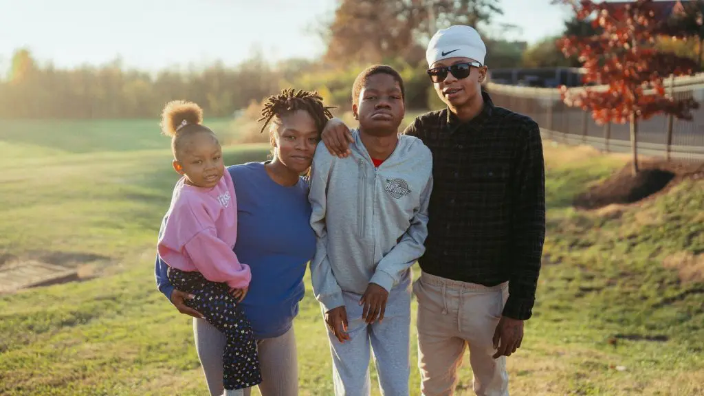 Family standing together outdoors in a grassy area at sunset, with one adult holding a young child.