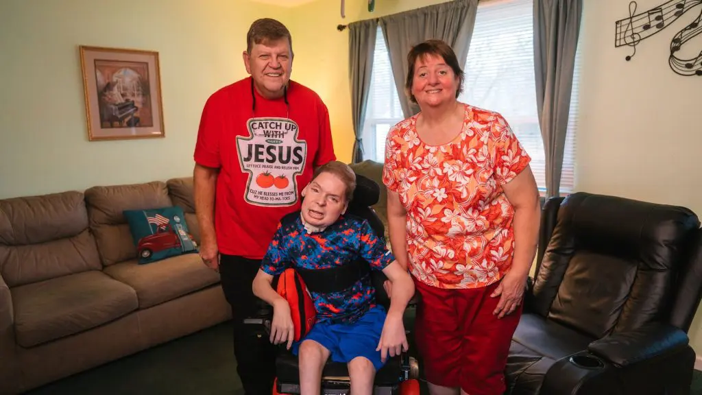 Three people smiling in a living room, with a young person seated in a wheelchair between two adults.