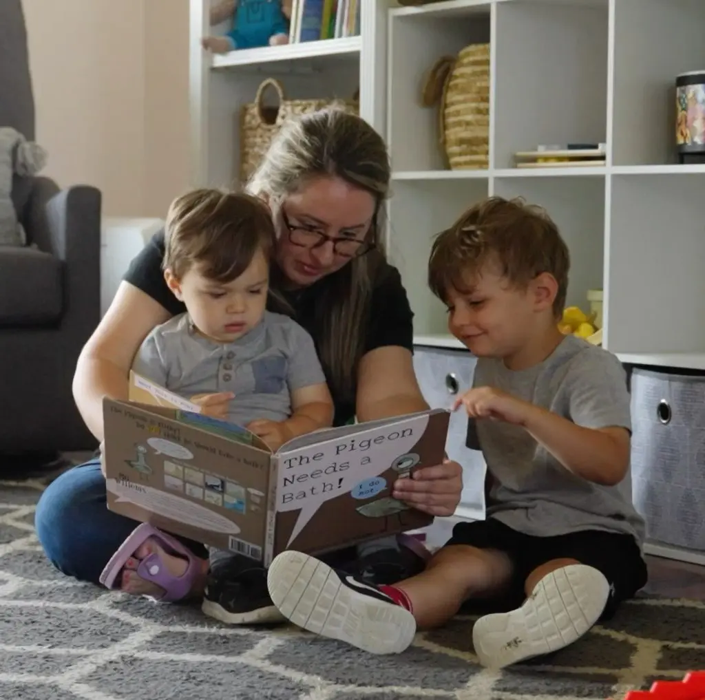 Adult sitting on the floor reading a picture book with two young children in a living room.