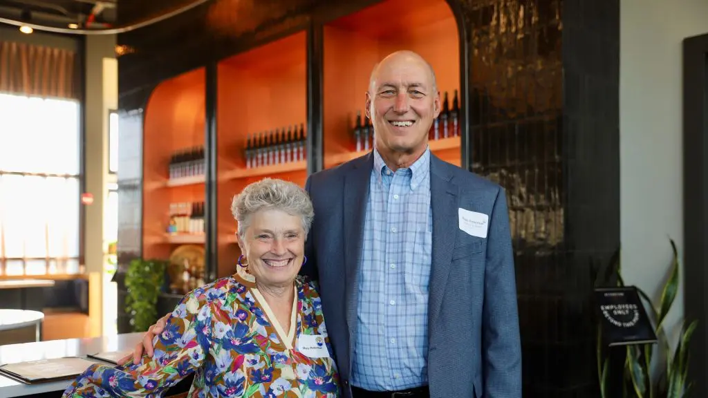 Two smiling adults posing together indoors in front of illuminated shelves displaying bottles.