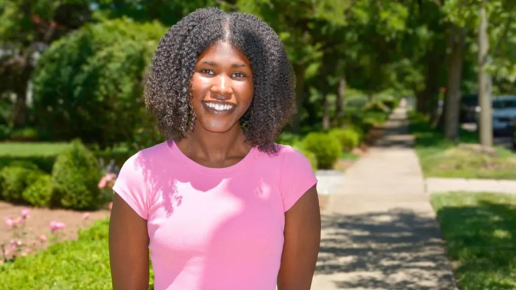 Smiling woman with curly hair wearing a pink shirt on a tree-lined sidewalk.
