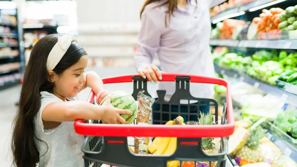 Child smiling while placing a small watermelon into a grocery cart filled with fruits and vegetables, next to an adult in a supermarket produce section.