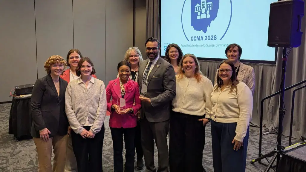 Group of nine adults posing at a conference event, with two people holding glass awards in front of a screen displaying “OCMA 2026 Future-Ready Leadership for Stronger Communities.”