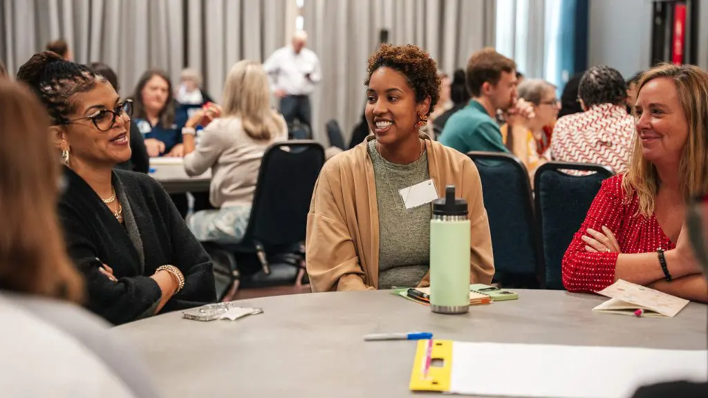 People seated around a round table engage in discussion during a partner meeting, with participants conversing in small groups throughout the room.
