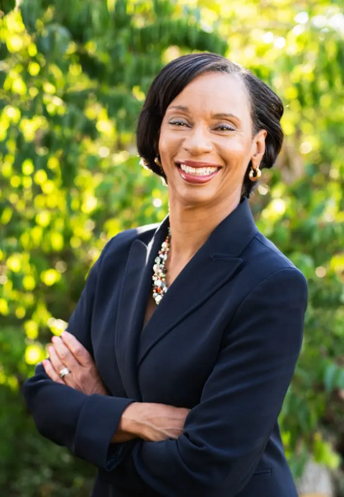 Professional headshot of a woman smiling with her arms crossed, standing outdoors in front of greenery.