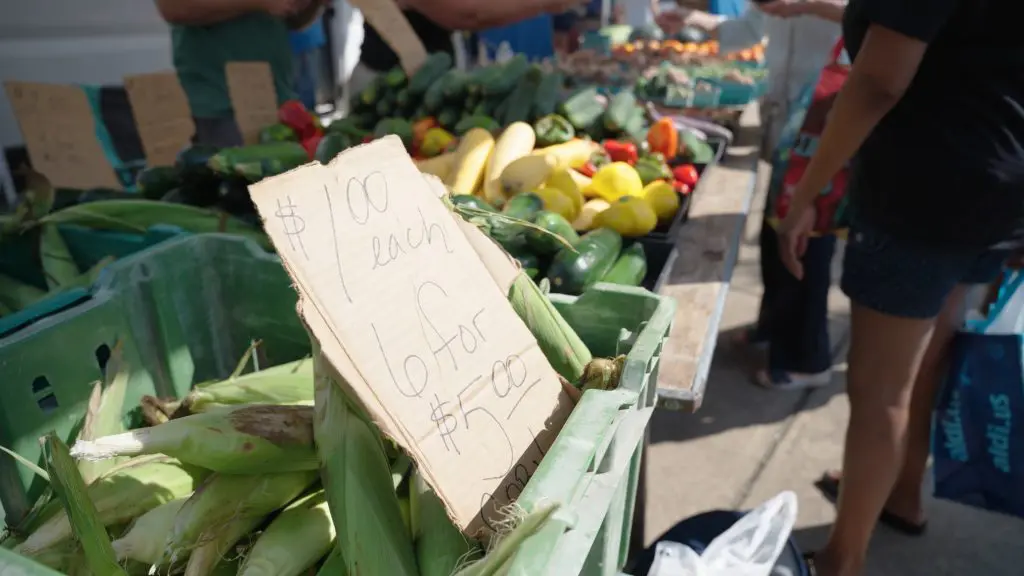 Fresh corn in green bins at a farmers market, with a handwritten sign advertising $1 per ear or six for $5. Other fresh vegetables and shoppers are visible in the background.