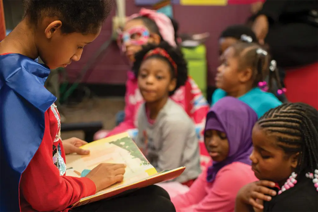 A child dressed as a superhero reads a book to a group of children seated together in a classroom.