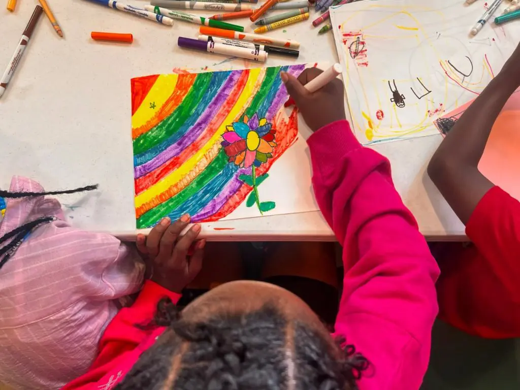 Children work on colorful drawings with markers at a table. One child colors a rainbow and flower while another draws nearby.