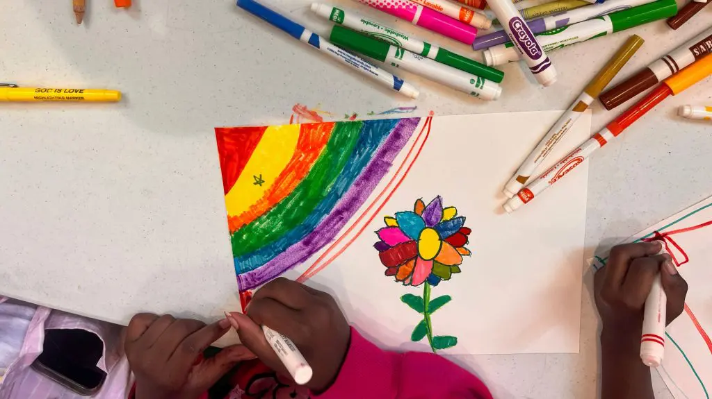 Overhead view of children creating colorful artwork with markers, including a rainbow and multicolored flower, on a table scattered with art supplies.