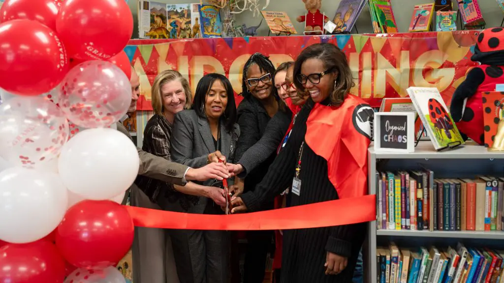 Group of people cutting a red ribbon at a library event, with red and white balloons, bookshelves, and a “Grand Opening” banner in the background.
