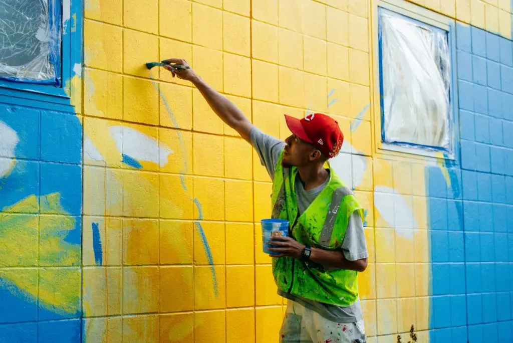 A person wearing a safety vest and red cap paints a yellow cinder block wall while holding a container of paint.