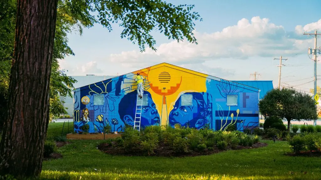 A person stands on a ladder painting a colorful mural on the front of a building surrounded by trees and landscaped gardens.