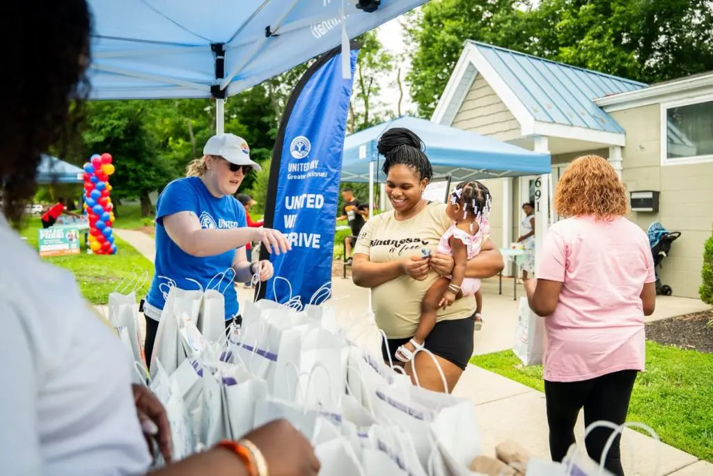 A volunteer hands out white gift bags to community members under a canopy at an outdoor neighborhood event.