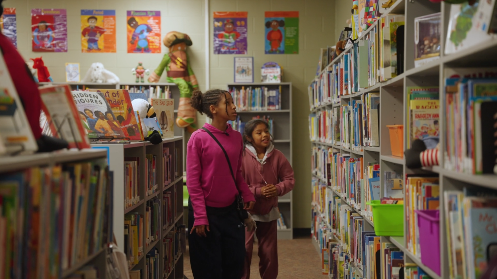 Two children browse books in the aisle of a children's library with colorful shelves, posters, and displays.