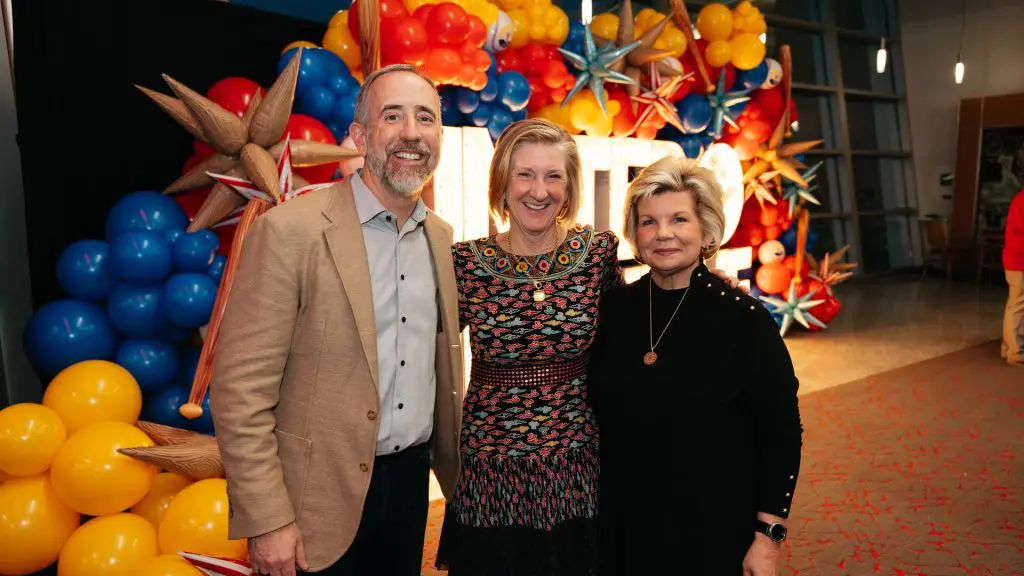Three people smiling and posing in front of a colorful balloon and star decoration display at an indoor event.