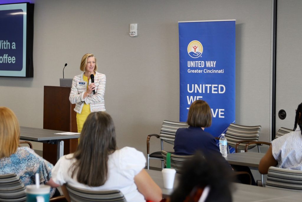 Woman speaking into a microphone at a meeting, standing near a podium and a United Way Greater Cincinnati banner while attendees listen.