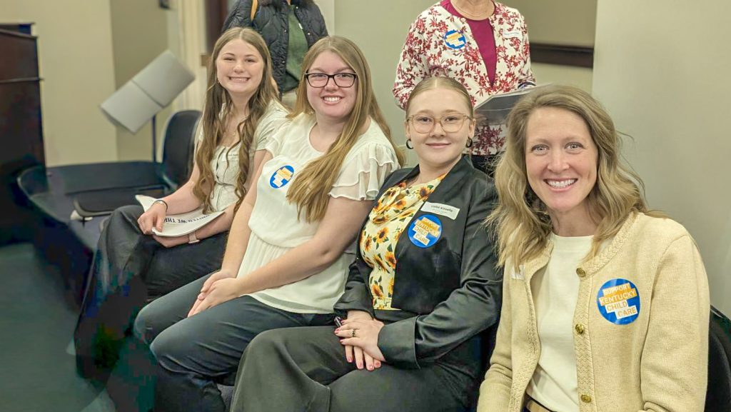 Four women seated at an indoor event wearing “Support Kentucky Child Care” stickers and smiling at the camera.