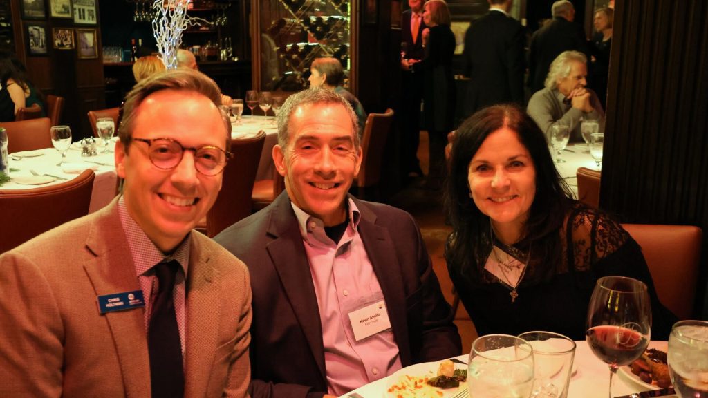 Three people smiling at a dinner event in a warmly lit restaurant, seated at a table with food and drinks.