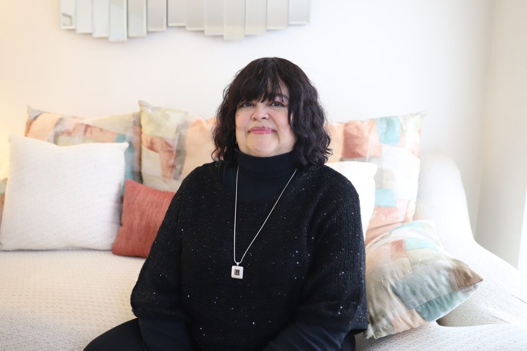A woman with dark, wavy hair sits on a couch, smiling. She wears a black turtleneck sweater and a silver pendant necklace. The couch has decorative pillows in neutral tones.
