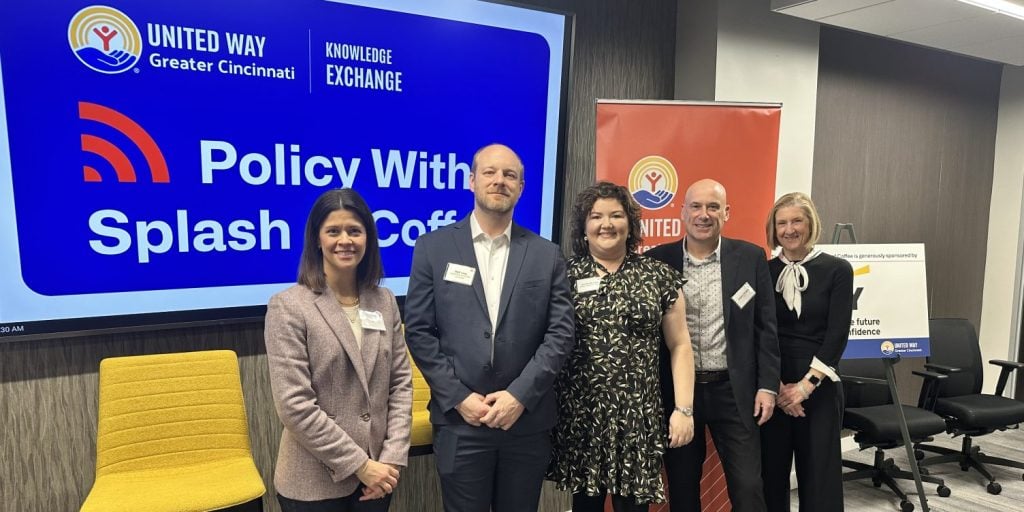 A group of five individuals pose for a photo at a United Way Greater Cincinnati event. They stand in front of a large screen displaying the event title "Policy With Splash Coffee" and the United Way and Knowledge Exchange logos. A red United Way banner and a sign with sponsorship information are visible in the background, along with yellow and black chairs in a modern conference room setting.