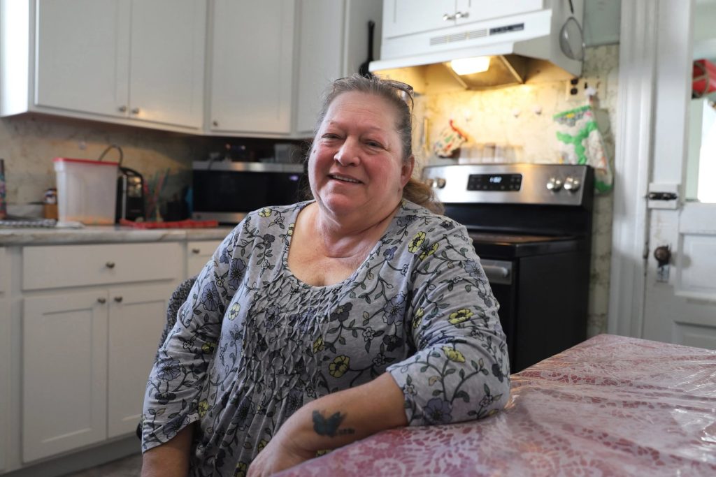 A woman in a floral blouse sits at a kitchen table, smiling. The kitchen has white cabinets, a stove, and a microwave in the background.